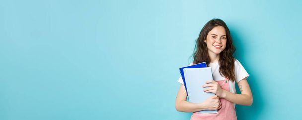 Милая улыбающаяся студентка с блокнотами и веселым видом на камеру, учится в колледже или университете, стоит на синем фоне. - Фото, изображение