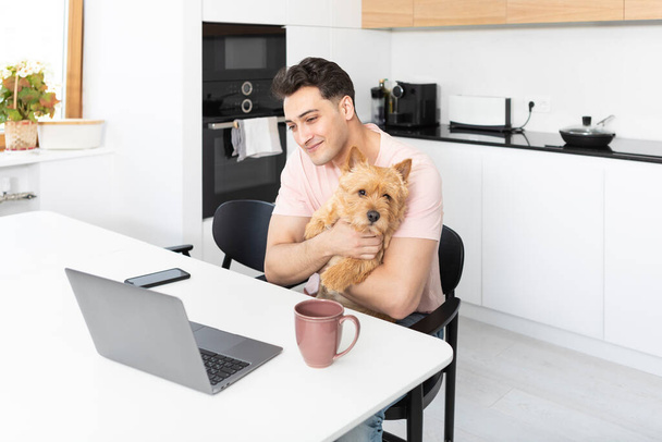 Happy handsome man hugs his pet dog Norwich Terrier and working on laptop at home - Foto, Imagen