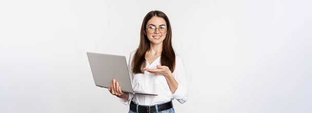 Ritratto di donna in occhiali che tiene il computer portatile, indica lo schermo, mostra il suo lavoro sul computer, in piedi su sfondo bianco. - Foto, immagini