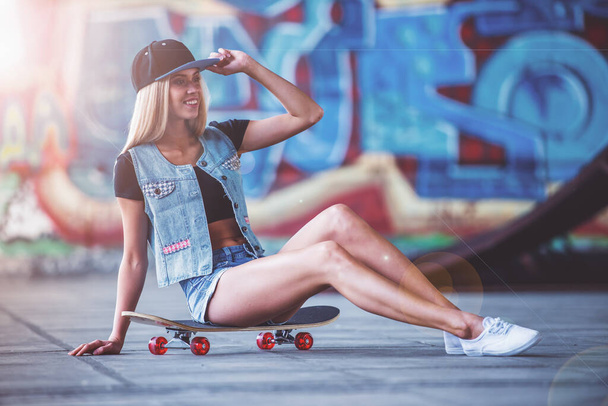 Beautiful blonde girl in cap is looking away and smiling while sitting on her skateboard in skate park - Photo, Image