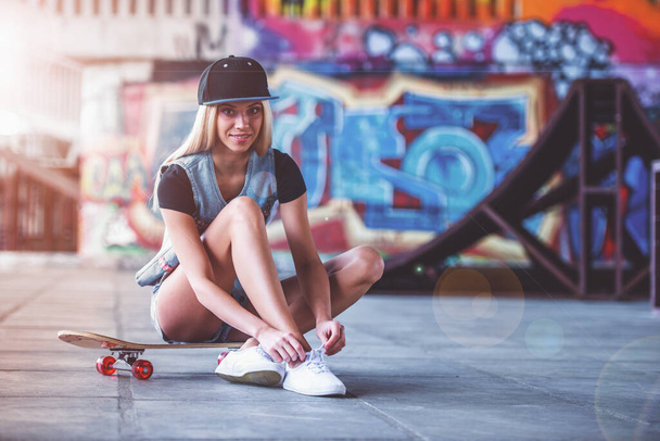 Beautiful blonde girl in cap is lacing her shoes, looking at camera and smiling while sitting on her skateboard in skate park - Photo, Image
