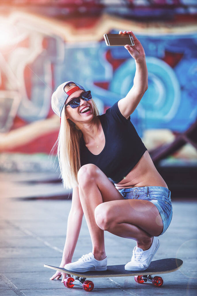 Hermosa chica rubia en gafas y gorra está haciendo selfie usando un teléfono inteligente y sonriendo mientras está de pie en su monopatín en el parque de skate
 - Foto, imagen