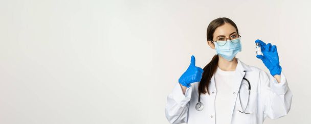 Close up portrait of young professional woman doctor, physician in medical face mask and glasses, showing vaccine and approve it, recommending get vaccinated from covid-19. - Foto, Imagem