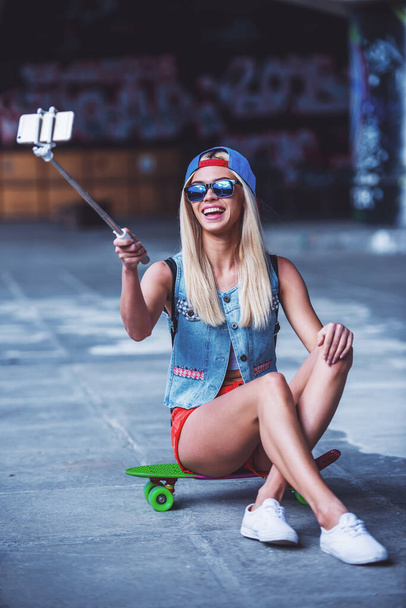 Beautiful blonde girl in cap and glasses is making selfie using a smart phone and smiling while sitting on her skateboard in skate park - Photo, Image