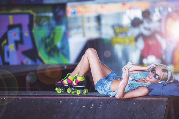 Вид сбоку красивой блондинки в наушниках и роликах, слушающей музыку через смартфон и улыбающейся, лежа в скейт-парке
 - Фото, изображение