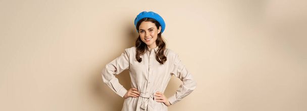 Portrait de femme tendance en trench-coat et chapeau français, souriant et heureux, posant dans une tenue élégante sur fond beige. - Photo, image