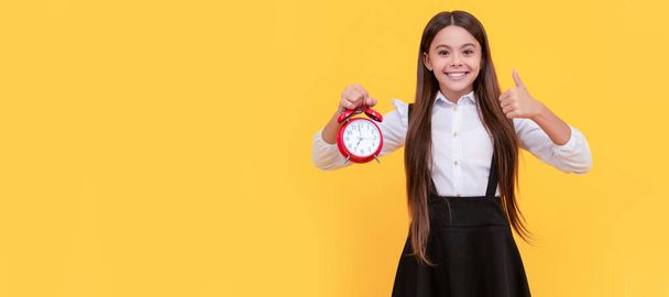 目覚まし時計で制服を着た幸せな10代の女の子時間と親指アップジェスチャー時間。時計アラーム、水平方向のポスターを持つ10代の子供。バナーヘッダー、コピースペース - 写真・画像