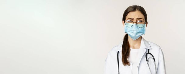 Сумна і похмура молода жінка-лікар в медичній масці для обличчя, почувається засмученою або стурбованою, хитається, стоїть на білому тлі. Копіювати простір
 - Фото, зображення