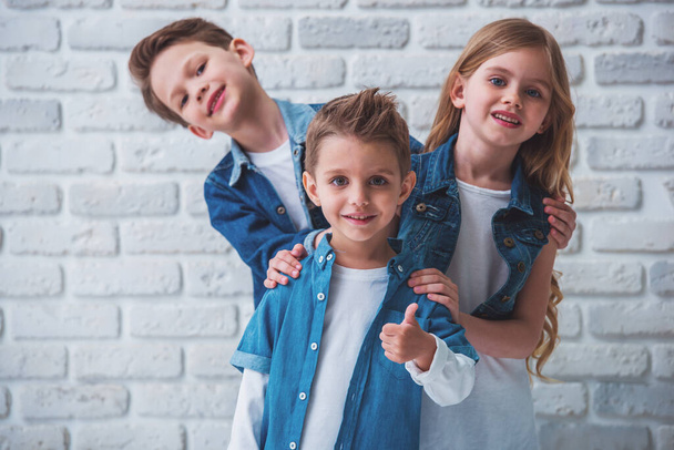 Cute little kids in stylish clothes are looking at camera and smiling, standing one by one against white brick wall. Front boy is showing Ok sign - Photo, image