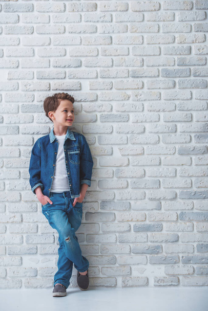 Ritratto a figura intera di un bambino elegante in jeans che distoglie lo sguardo e sorride, in piedi con le mani in tasca contro il muro di mattoni bianchi
 - Foto, immagini