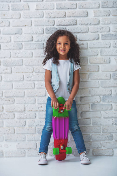 可憐なアフロ アメリカン女の子のカメラ目線や笑顔、白いレンガの壁に対して彼女のスケート ボードに立って - 写真・画像