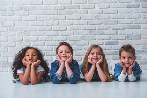 Cute little kids in stylish jeans clothes are leaning on their hands, looking at camera and smiling, lying on the floor against white brick wall - Photo, Image