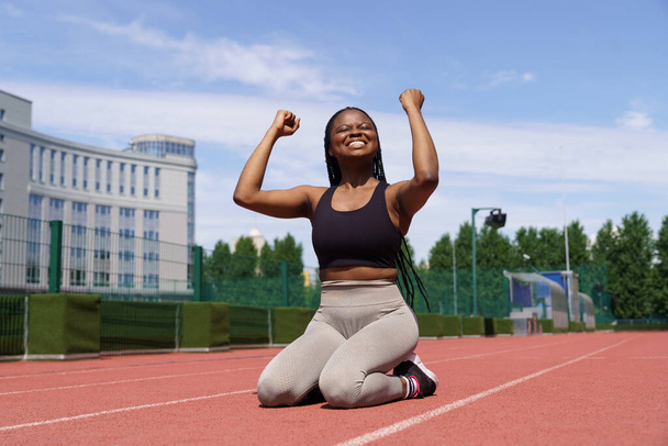 Cheerful woman raises hands sitting on red track at sports ground against modern building and city park. African American athlete feels happy after successful running long distance on sunny summer day - Foto, imagen