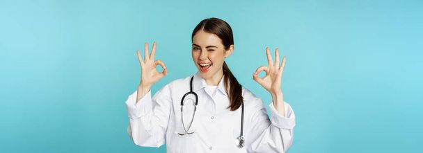 Усміхнена жінка-лікар, професійний медичний працівник, показує добре, добре знак схвалення, рекомендує клініку, карантинну кампанію, бірюзовий фон
. - Фото, зображення