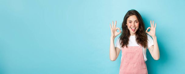 Portrait of excited brunette girl with curly hairstyle, showing okay signs, praise perfect choice, recommend good thing, give OK sign, standing over blue background. - Photo, Image