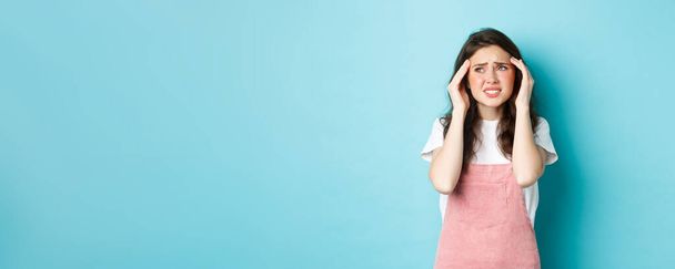 Frau mit schmerzhafter Migräne, Berührung des Kopfes und Stirnrunzeln, Blick in die linke obere Ecke, Kopfschmerzen, vor blauem Hintergrund stehend. - Foto, Bild