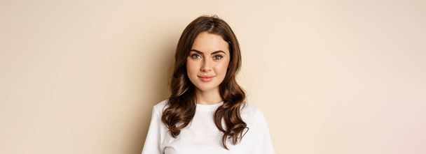 People and emotions. Close up of confident smiling young woman, looking at camera, standing in casual relaxed pose, wearing white t-shirt, beige studio background. - Foto, Imagen