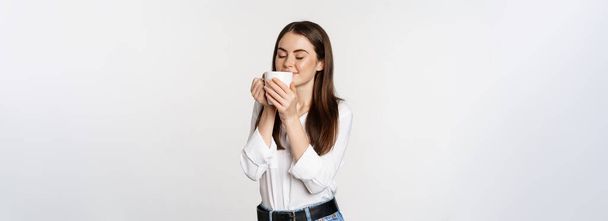 Lächelnde Bürofrau trinkt Kaffee, trinkt zufrieden aus Becher, genießt, steht vor weißem Hintergrund. - Foto, Bild