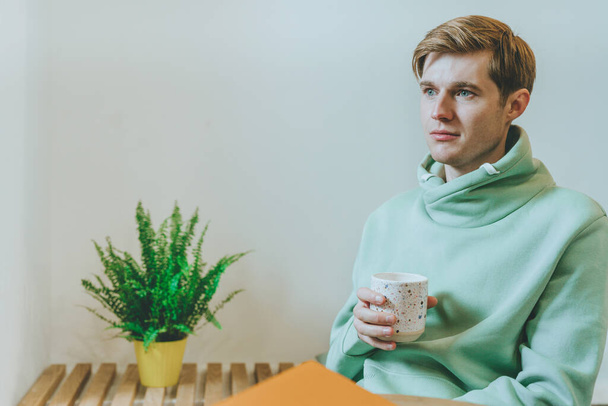 Kafede oturan düşünceli genç bir adam bir fincan kahveyle pencereye bakıyor. Seçici odak. - Fotoğraf, Görsel