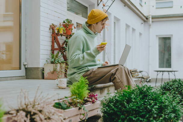 Фрилансер молодой человек, работающий на ноутбуке, используя смартфон, сидящий на лестнице на улице. Селективный фокус. - Фото, изображение