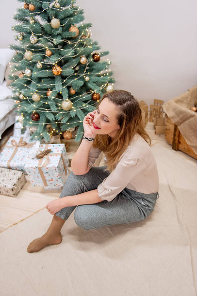 Stylish, modern millennial woman sits near Artificial Christmas tree, wooden box, chest with glitter balls. Festive eco-friendly interior, with festive gift boxes by the bed. - Φωτογραφία, εικόνα