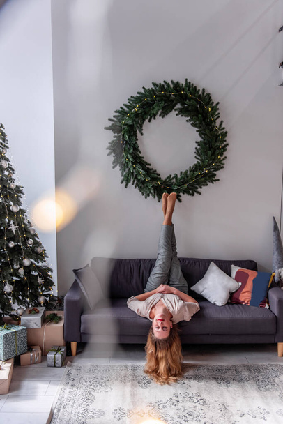 Millennial woman with red lipstick having fun lying upside down on gray sofa near the Christmas tree. Crazy New Year's Eve. Relax in the morning after the party. Scandinavian style in interior design - Photo, image