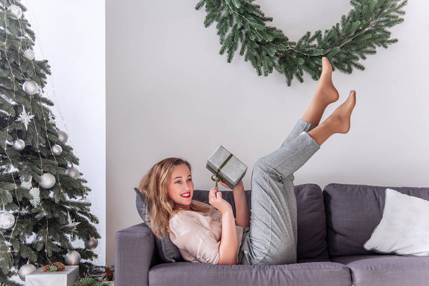 Millennial woman with red lipstick in pin-up style, holding a gift in hands, legs raised up on a gray sofa near the Christmas tree, wreath. Crazy New Year's Eve. Relax in the morning after the party. - Фото, зображення