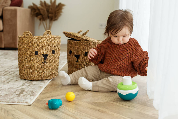 Toddler child is playing with toys on floor in room. Childhood, kindergarten and development concept. Brown sweater - Photo, image