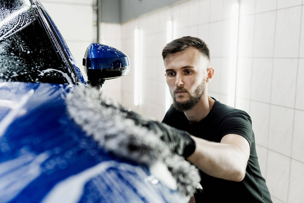 Hand washing with microfiber glove with foam car body in garage. Car washer doing manual foam washing in auto detailing service - Fotó, kép