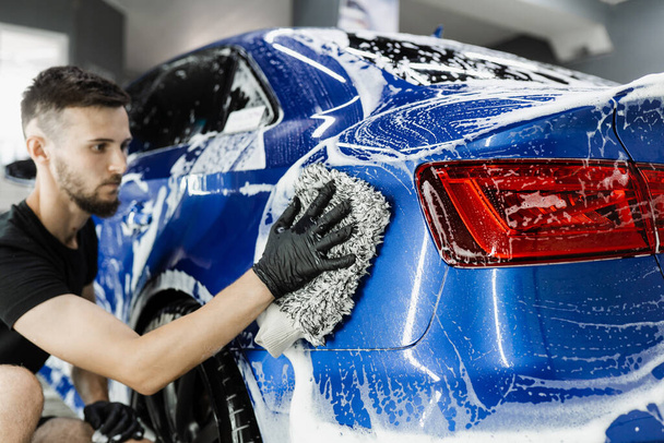 Autowaschanlage beim manuellen Schaumwaschen im Auto-Detaillierungsservice. Händewaschen mit Mikrofaserhandschuh mit Schaumstoffkarosserie in der Garage - Foto, Bild