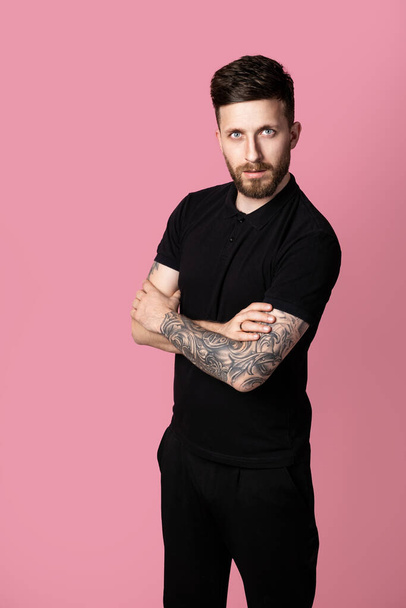 Fashionable bearded handsome hipster man with stylish haircut and in fashionable casual clothes total black standing with crossing hands isolated on pink studio background  - Fotó, kép