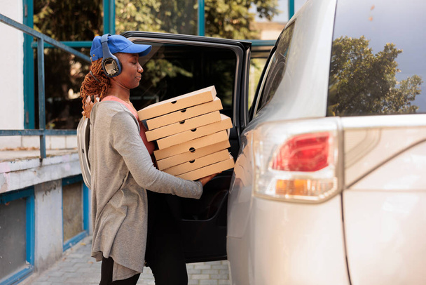 Office lunch delivery service worker delivering pizza by car, side view. African american courier in headphones holding fastfood boxes stack, standing near company building outdoors - Photo, Image