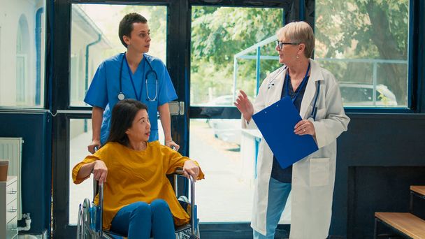 Infirmière aidant une femme asiatique ayant une déficience physique à consulter un médecin. Utilisateur de fauteuil roulant souffrant d'une déficience chronique dans la salle d'attente dans le hall de la clinique. - Photo, image