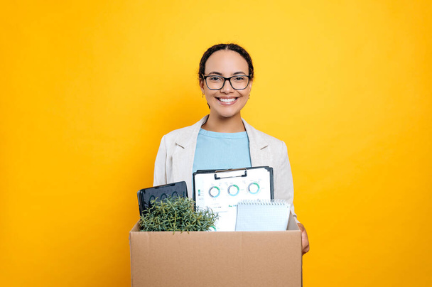 New job. Excited positive lovely mixed race female employee, newcomer, in stylish elegant clothes, holding cardboard box, stand on isolated orange background, looking at camera, smiling friendly - Photo, Image