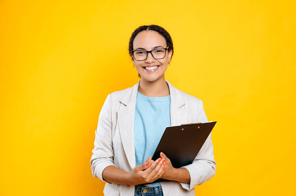 Happy positive lovely mixed race female employee, real estate agent, recruitment specialist, hr manager, holding documents, stands on isolated orange background, looking at camera, smiling friendly - Fotó, kép