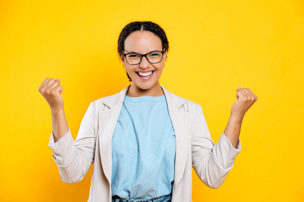 Win, success. Overjoyed mixed race business woman with glasses, rejoices at good news, victory, deal, gesturing with fists, stands on isolated orange background, looks at camera, smiling happily - Foto, afbeelding
