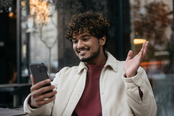Sorrindo homem indiano elegante usando telefone celular acenando mão, comunicação on-line na rua. Feliz sucesso asiático influenciador gravação de vídeo ao ar livre  - Foto, Imagem