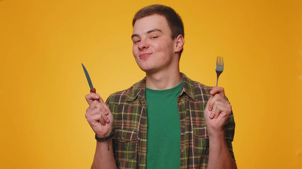 Ready to eat. Hungry teenager adult boy waiting for serving dinner dishes with cutlery will appreciate delicious restaurant meal. Young man guy with fork and knife isolated on yellow studio background - Photo, Image
