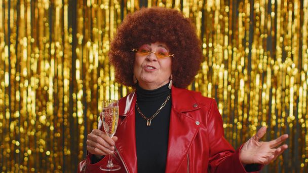 Funny woman in red jacket listening music, smiling dancing to disco party music, drinking champagne, having fun. Senior grandmother celebrating winning, birthday, xmas isolated on shiny background - Photo, Image
