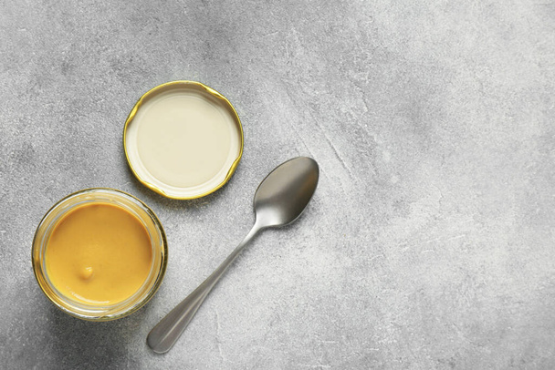 Spicy mustard in glass jar and spoon on grey table, flat lay. Space for text - Φωτογραφία, εικόνα