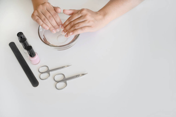Hands of woman are in bath with warm water with useful substances. Nail file, scissors and bottles of varnish are next on table, copy space. Concept of nail and cuticle and hand skin care - Foto, imagen