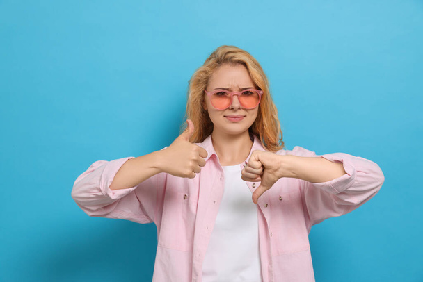 Conflicted young woman showing thumbs up and down gestures on light blue background - Foto, immagini