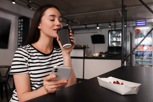Pansiyonun yemek odasındaki masada elinde bir fincan kahve ve akıllı telefonla mutlu genç bir kadın. - Fotoğraf, Görsel