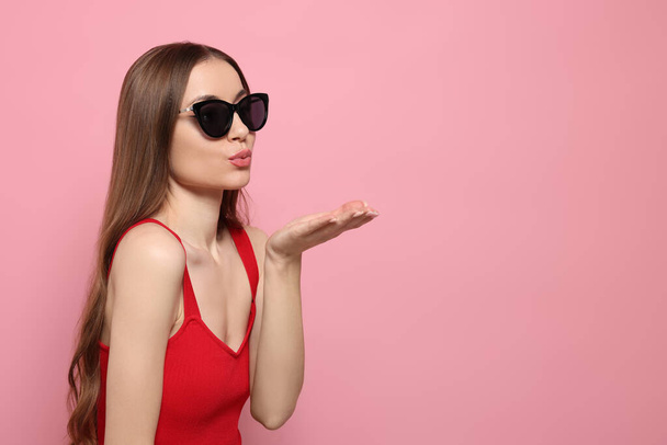 Beautiful young woman with sunglasses blowing kiss on pink background, space for text - Фото, изображение