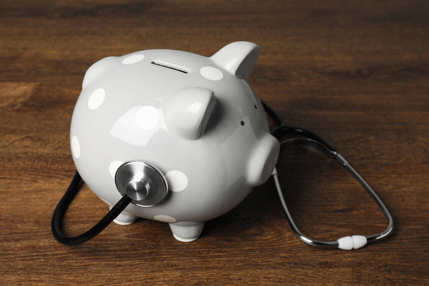 Light gray ceramic piggy bank and stethoscope on wooden table. Medical insurance - Φωτογραφία, εικόνα