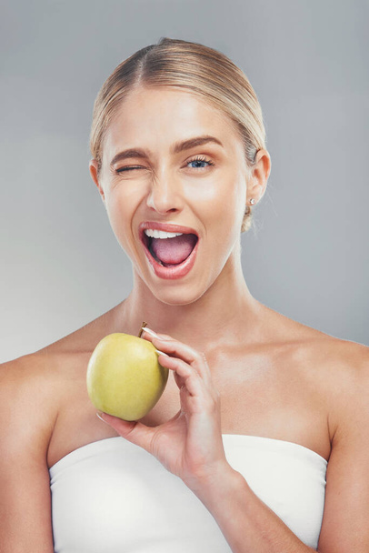 Woman, health and wink portrait with apple for fiber diet, nutrition and wellness lifestyle. Fruit, healthy and digestion dieting marketing with happy model girl on gray studio background - Foto, imagen