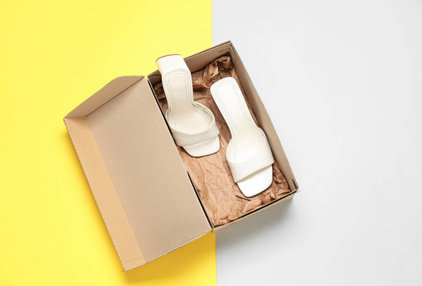 Caja de cartón con sandalias de tacón blanco sobre fondo de color - Foto, Imagen