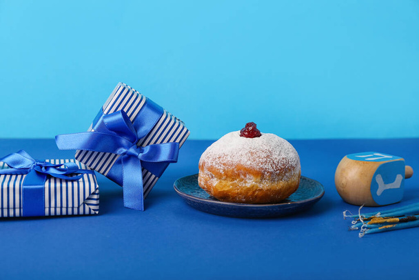 Assiette avec beignet savoureux, dreidel, bougies et cadeaux pour la célébration Hannukah sur fond bleu - Photo, image