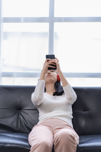 Krásná asijská žena nosí sluchátka, poslouchá hudbu, používá svůj smartphone, zatímco chlazení na pohovce v obývacím pokoji doma na dovolenou. - Fotografie, Obrázek