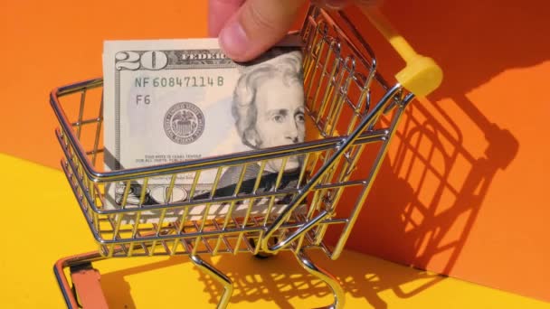 Hand toevoegen 20 dollar bankbiljet Geld in speelgoed supermarkt trolley winkelwagen op gele achtergrond. Verkoopmand met US dollar biljetten. Minimaal leefbaar loon Concept: lening, beleggingspensioen - Video